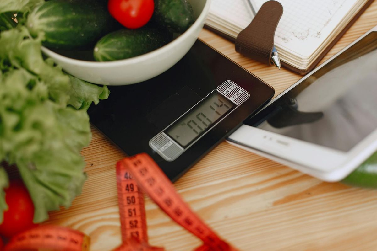 A bowl of fresh vegetables on a kitchen scale alongside measurement tools — the working setup of someone tracking calories.