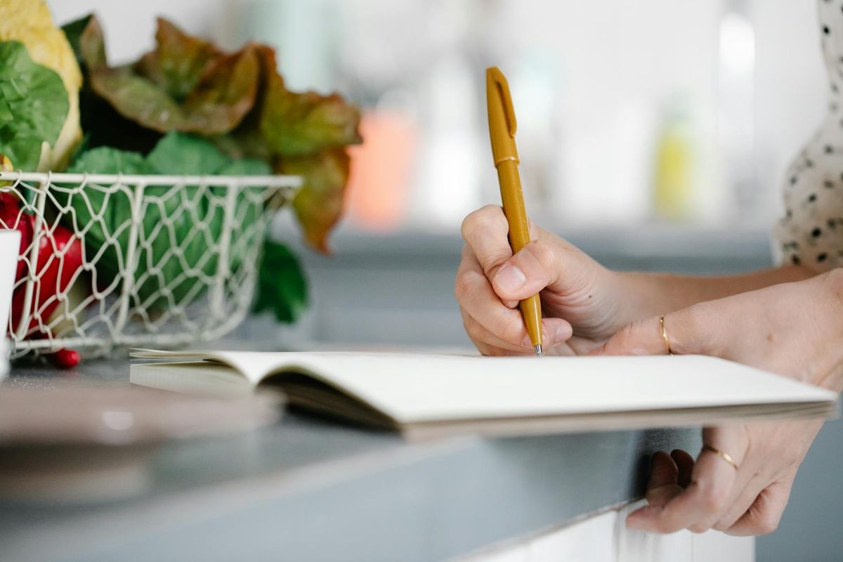 A woman writing in a notebook on a kitchen counter beside a basket of fresh vegetables.