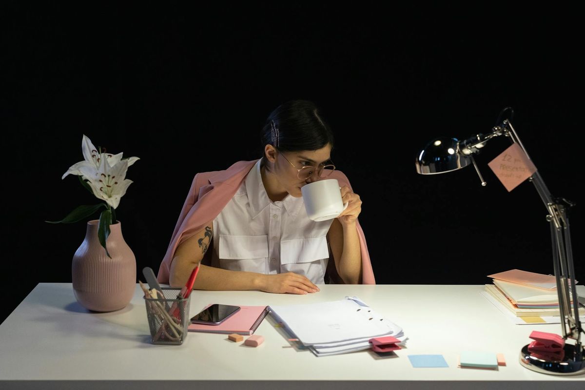 A young woman in an office setting drinking coffee, surrounded by work items and a vase of flowers.